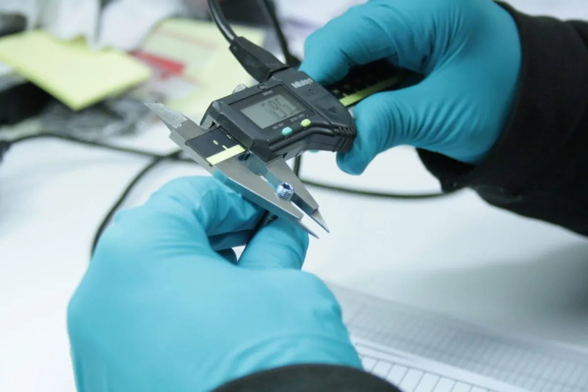 Quality inspector in nitrile gloves measuring a small machined part with a Mitutoyo digital caliper at Brassland